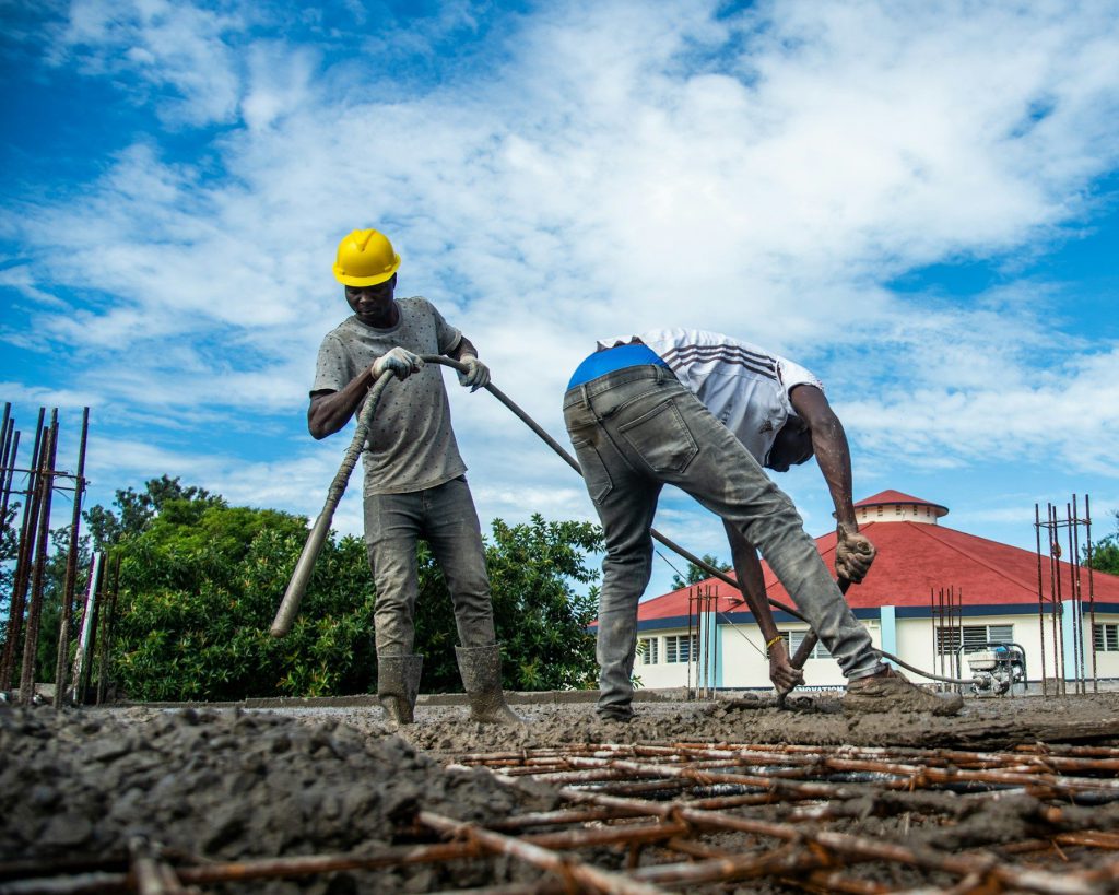 Hai công nhân, một người đội mũ bảo hộ vàng, dùng cây gạt san bê tông tươi trên lưới thép sàn ngoài trời, phía sau là một ngôi nhà mái đỏ và bầu trời xanh